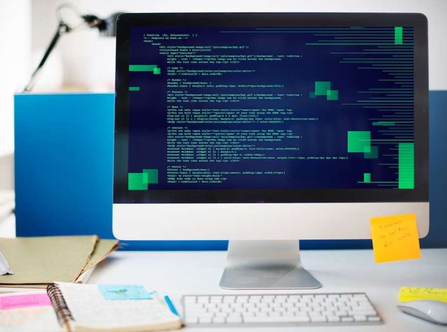 computer screen with coding on it, yellow note, keyboard, computer mouse, and notes on the table